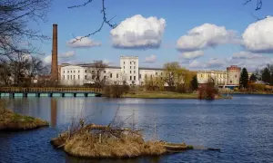 Kazimierza Wielka atrakcje: nad jeziorem fabryka z ceglanym kominem i wieżą, mostek, błękitne niebo z chmurami.