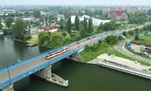 Tram z czerwonymi pasami jedzie przez most nad rzeką, w tle widać miasto i tereny przemysłowe, jak na plaży Stogi.