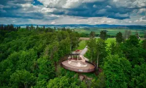 Widok na taras widokowy w otoczeniu lasu, z panoramą pól i gór w tle. Jakby to było jezioro wśród gór stołowych.