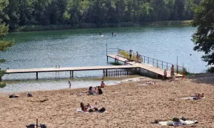Słoneczna borówno plaża nad jeziorem, ludzie odpoczywają na piasku i w wodzie, niektórzy pływają na desce SUP.