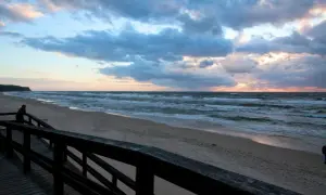 Zachód słońca nad morzem, plaża w Wiśle. Drewniany pomost prowadzi do piaszczystego brzegu, gdzie fale rozbijają się o brzeg.
