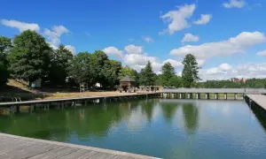 Mrągowo plaża z drewnianym molo i zieloną wodą, otoczona drzewami pod błękitnym niebem z chmurami.