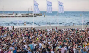 Tłumy ludzi na plaży w Sopocie, cieszące się letnimi imprezami. W tle morze i port.
