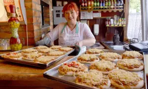 Uśmiechnięta kobieta z rudymi włosami przygotowuje pizze w najstarszej pizzerii w Polsce. Na tacach leżą gotowe do pieczenia placki z serem.
