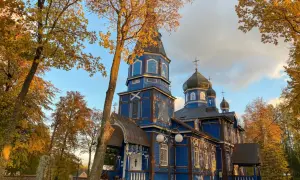 Niebieska cerkiew w jesiennym otoczeniu, z liśćmi w barwach złota i brązu. W oddali widać drzewa, a na pierwszym planie kamienny obelisk.