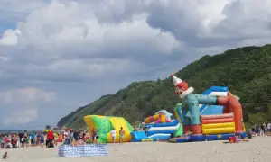 Wesołe miasteczko na plaży w Międzyzdrojach z dmuchanym klaunem i zjeżdżalniami. Ludzie odpoczywają, a w tle widać las i morze.