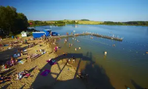 Słoneczny dzień nad zalewem w Siamoszycach. Ludzie odpoczywają na plaży i kąpią się w wodzie. Sprawdź cennik atrakcji!