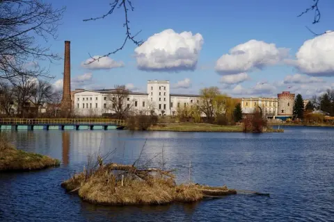 Kazimierza Wielka atrakcje: nad jeziorem fabryka z ceglanym kominem i wieżą, mostek, błękitne niebo z chmurami.