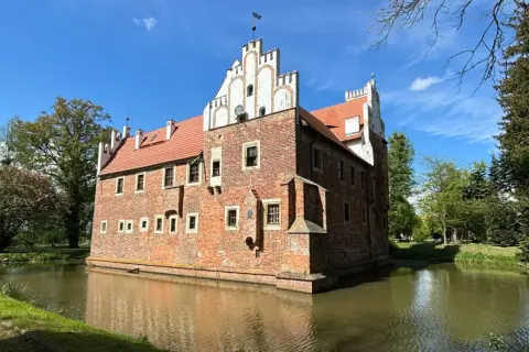 Zamek w Wojnowicach otoczony fosą, z ceglaną fasadą i czerwoną dachówką, pod błękitnym niebem.