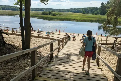 Dziewczynka schodzi drewnianymi schodkami na piaszczystą plażę nad spokojne stawy. W oddali widać ludzi kąpiących się i odpoczywających.