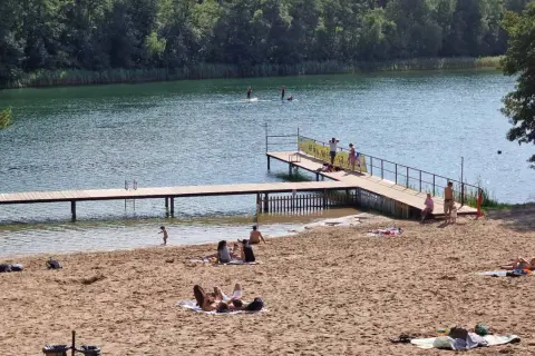 Słoneczna borówno plaża nad jeziorem, ludzie odpoczywają na piasku i w wodzie, niektórzy pływają na desce SUP.