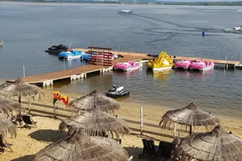 Słoneczna plaża Skorochów z parasolami, leżakami i kolorowymi łódkami wodnymi przy drewnianym pomoście.