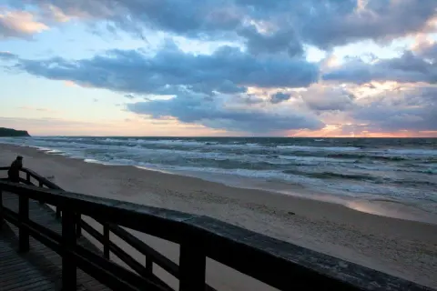 Zachód słońca nad morzem, plaża w Wiśle. Drewniany pomost prowadzi do piaszczystego brzegu, gdzie fale rozbijają się o brzeg.
