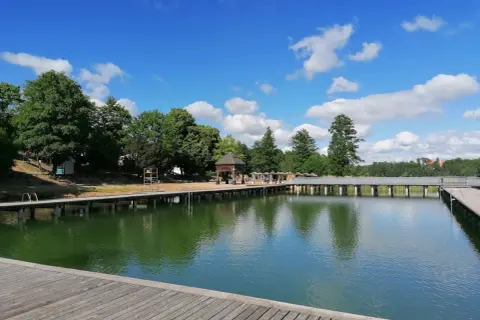 Mrągowo plaża z drewnianym molo i zieloną wodą, otoczona drzewami pod błękitnym niebem z chmurami.