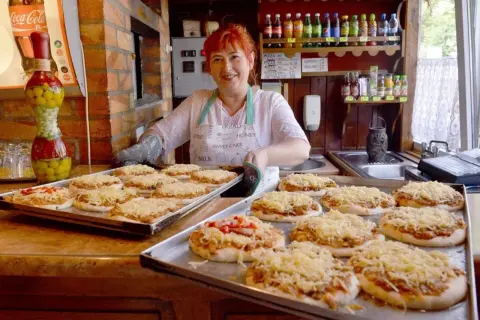 Uśmiechnięta kobieta z rudymi włosami przygotowuje pizze w najstarszej pizzerii w Polsce. Na tacach leżą gotowe do pieczenia placki z serem.