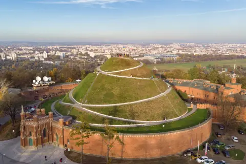 Kopiec Kościuszki, zielona kopuła z krętymi ścieżkami, góruje nad ceglanymi fortami i rozległym, **starym miastem w Krakowie**.