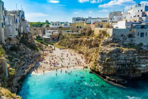 Słoneczne bari plaże z turkusową wodą, skalistymi klifami i białymi budynkami. Ludzie odpoczywają na piasku i pływają.