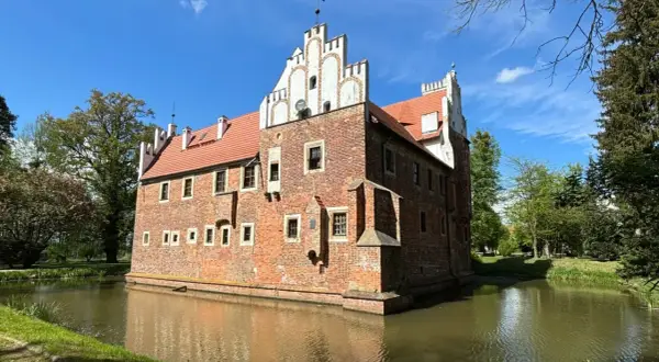Zamek w Wojnowicach otoczony fosą, z ceglaną fasadą i czerwoną dachówką, pod błękitnym niebem.