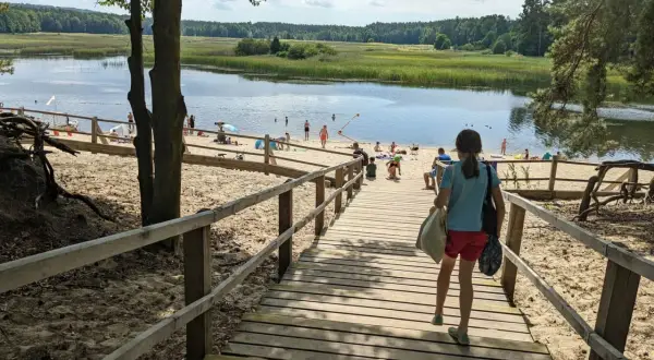 Dziewczynka schodzi drewnianymi schodkami na piaszczystą plażę nad spokojne stawy. W oddali widać ludzi kąpiących się i odpoczywających.