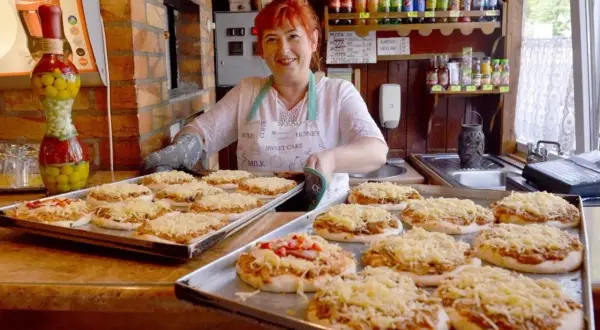 Uśmiechnięta kobieta z rudymi włosami przygotowuje pizze w najstarszej pizzerii w Polsce. Na tacach leżą gotowe do pieczenia placki z serem.