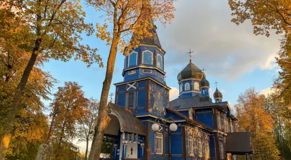 Niebieska cerkiew w jesiennym otoczeniu, z liśćmi w barwach złota i brązu. W oddali widać drzewa, a na pierwszym planie kamienny obelisk.