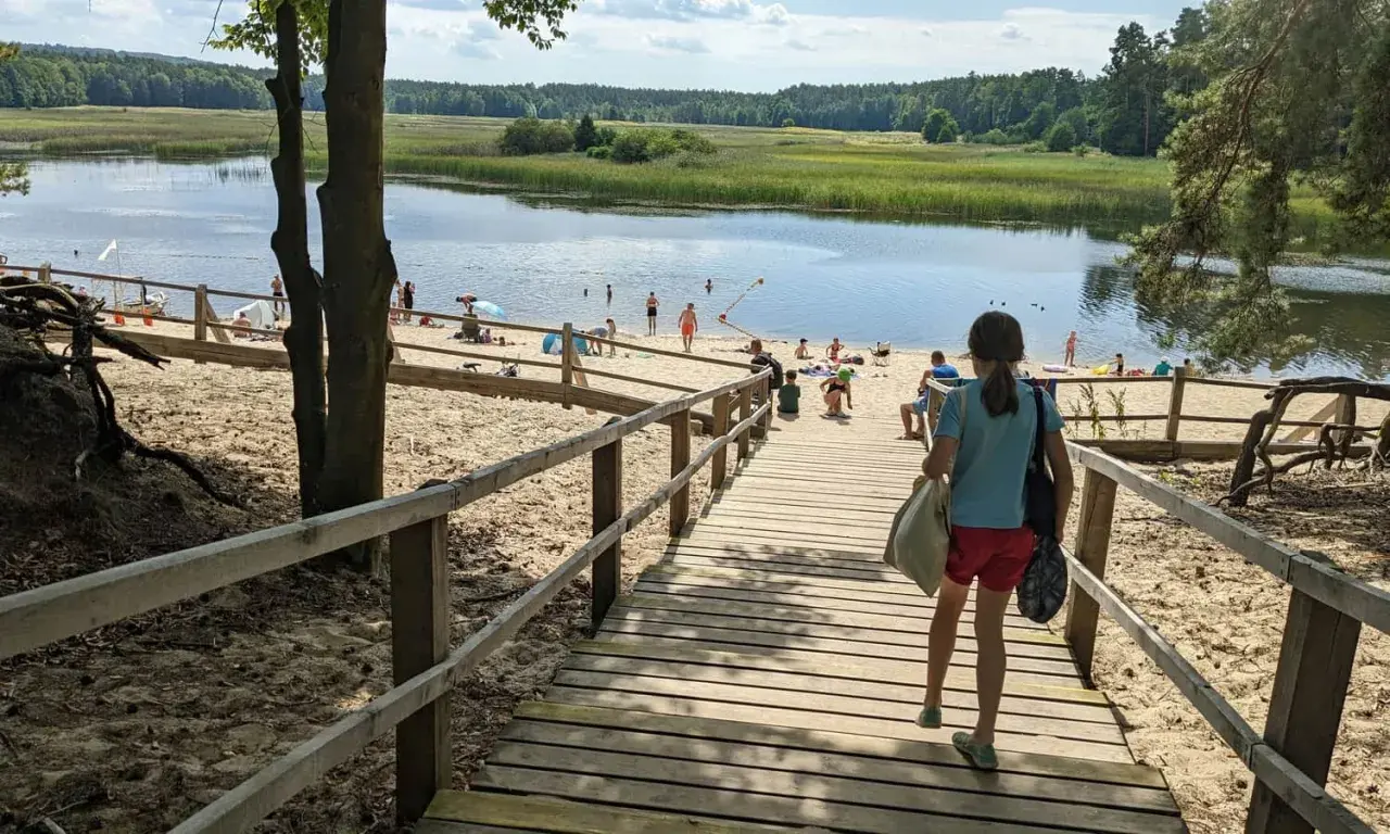 Dziewczynka schodzi drewnianymi schodkami na piaszczystą plażę nad spokojne stawy. W oddali widać ludzi kąpiących się i odpoczywających.