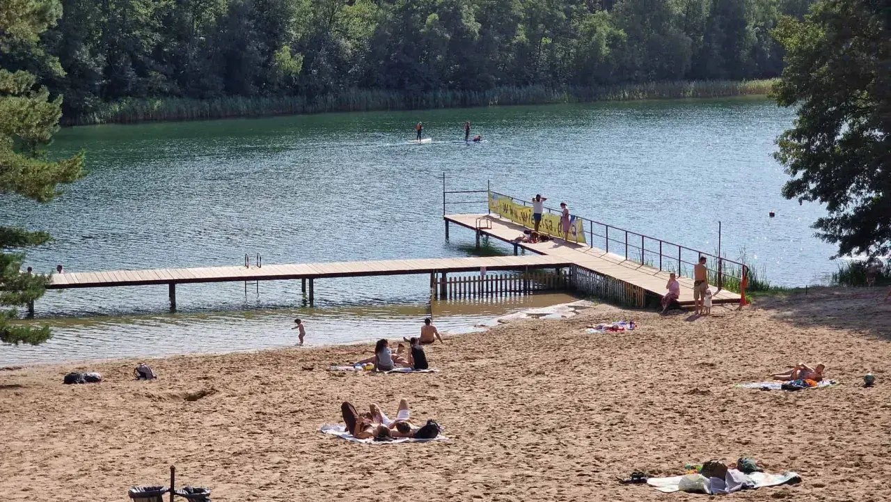 Słoneczna borówno plaża nad jeziorem, ludzie odpoczywają na piasku i w wodzie, niektórzy pływają na desce SUP.