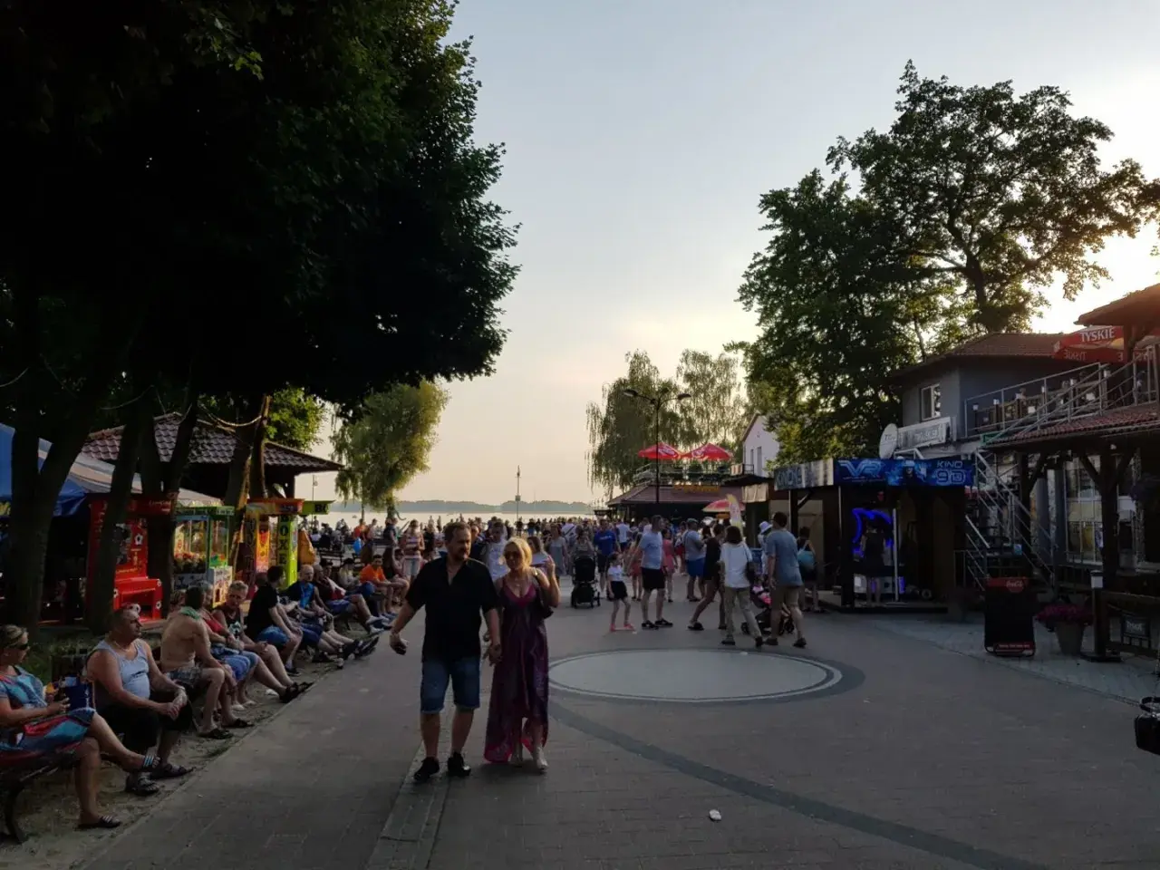 Tłum ludzi spaceruje promenadą w Skorzęcinie, korzystając z licznych atrakcji i letniej atmosfery nad jeziorem.