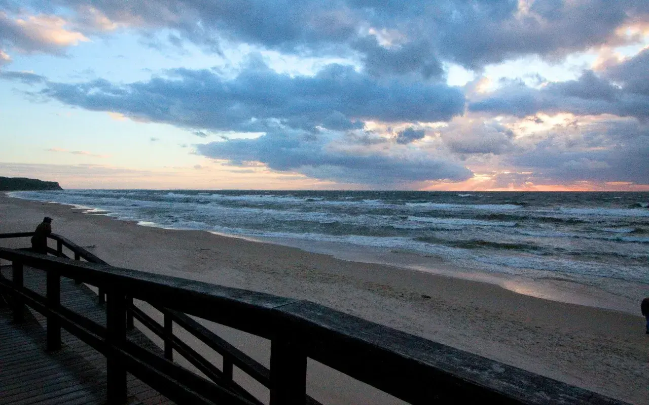 Zachód słońca nad morzem, plaża w Wiśle. Drewniany pomost prowadzi do piaszczystego brzegu, gdzie fale rozbijają się o brzeg.