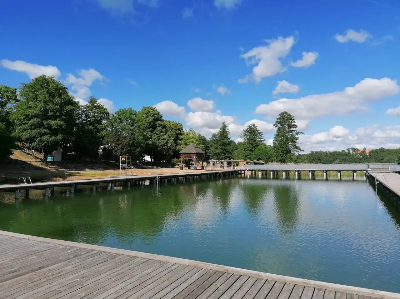 Mrągowo plaża z drewnianym molo i zieloną wodą, otoczona drzewami pod błękitnym niebem z chmurami.