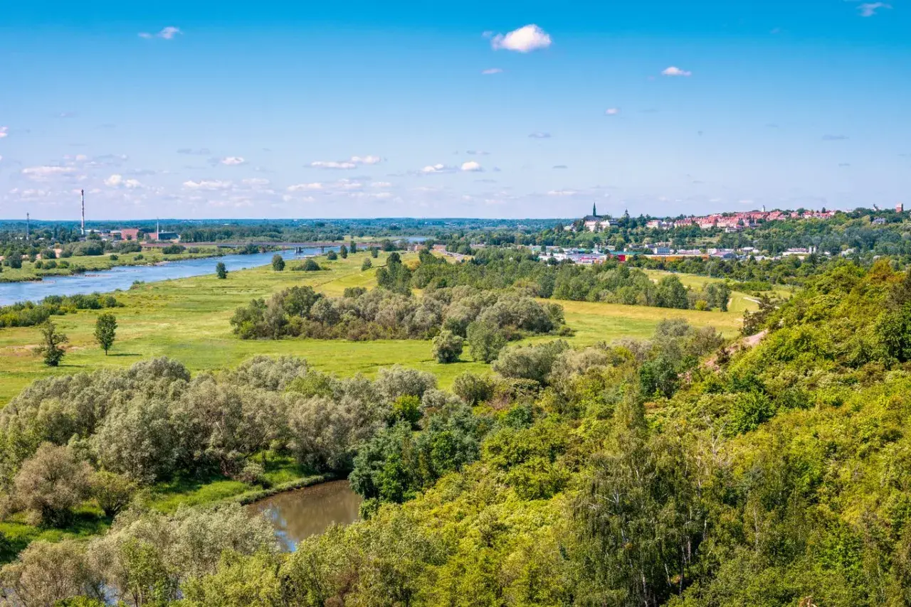 Krajobraz z rzeką, zielonymi wzgórzami porośniętymi drzewami i miastem w oddali.