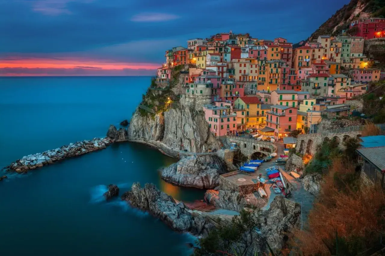 Kolorowe domy Cinque Terre w **Włoszech** wznoszą się nad skalistym wybrzeżem. **Morze** Liguryjskie lśni w blasku zachodzącego słońca.