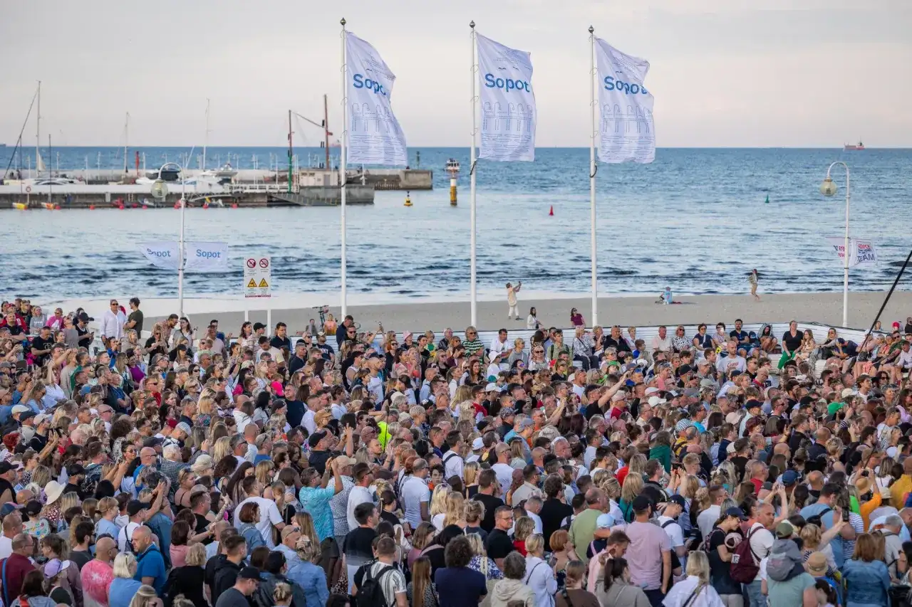 Tłumy ludzi na plaży w Sopocie, cieszące się letnimi imprezami. W tle morze i port.