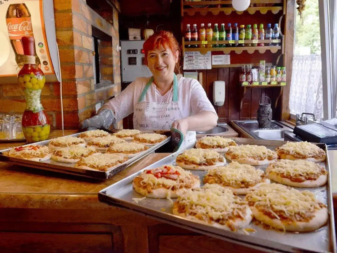 Uśmiechnięta kobieta z rudymi włosami przygotowuje pizze w najstarszej pizzerii w Polsce. Na tacach leżą gotowe do pieczenia placki z serem.