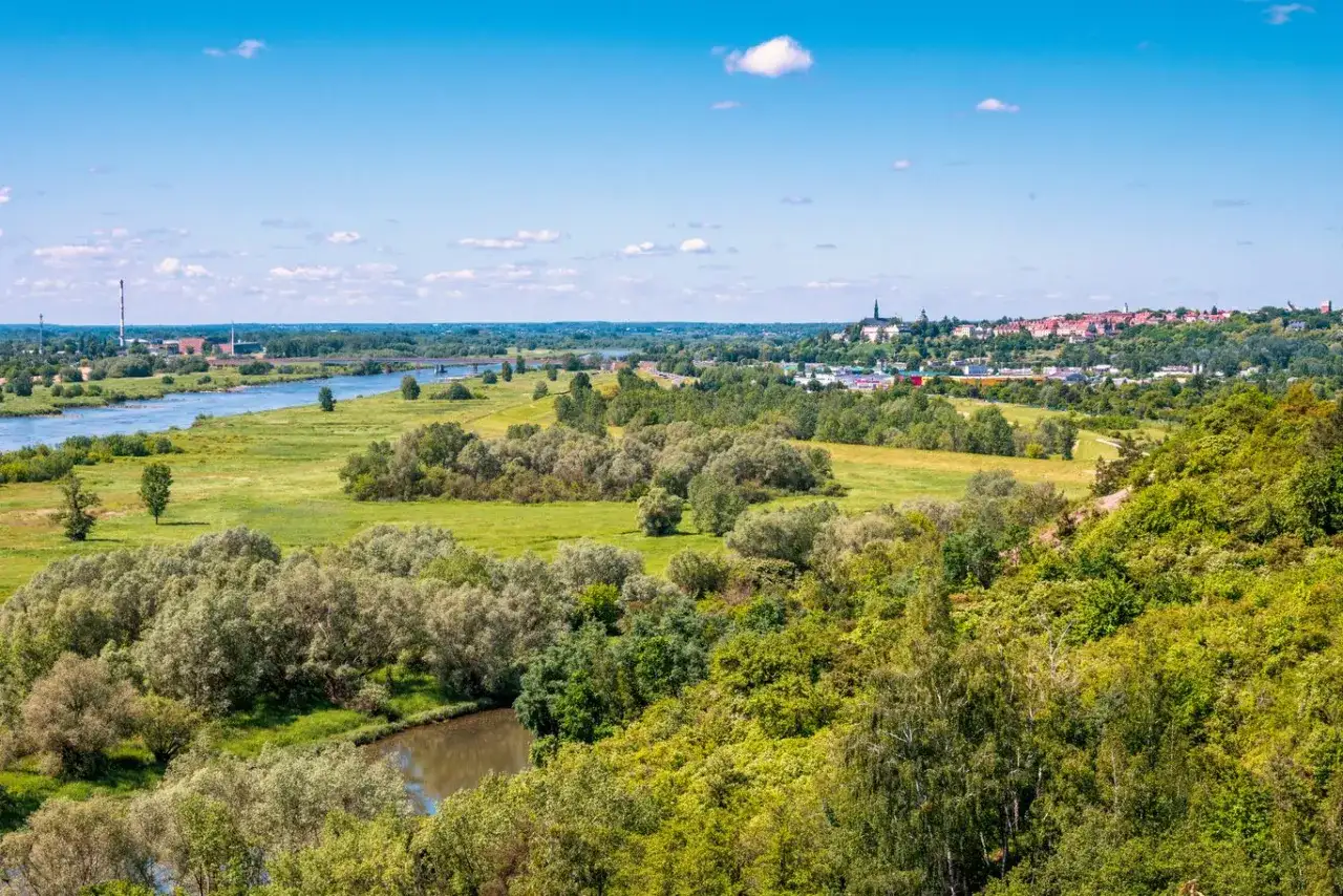 Krajobraz z rzeką, zielonymi wzgórzami porośniętymi drzewami i miastem w oddali.