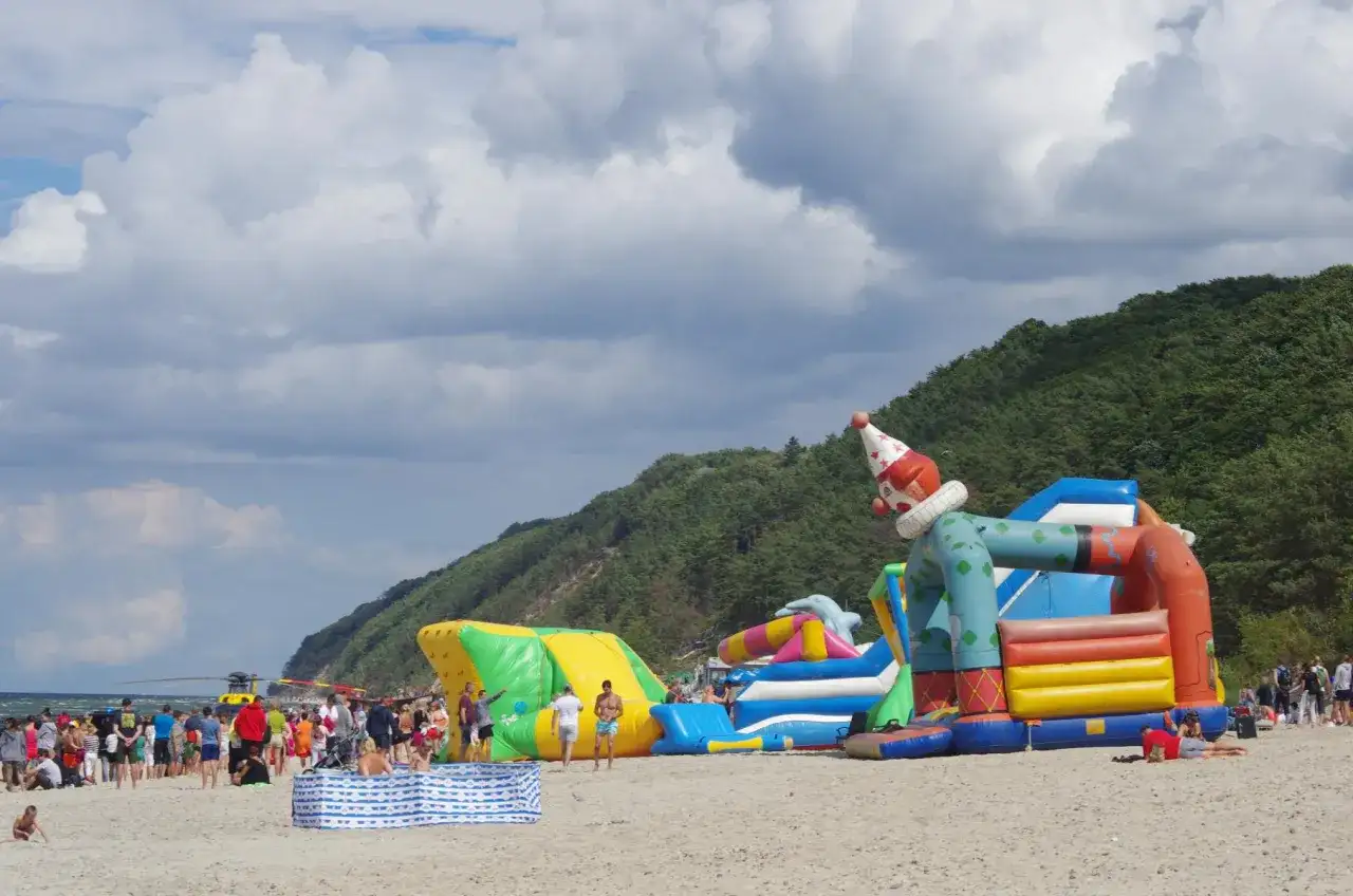 Wesołe miasteczko na plaży w Międzyzdrojach z dmuchanym klaunem i zjeżdżalniami. Ludzie odpoczywają, a w tle widać las i morze.