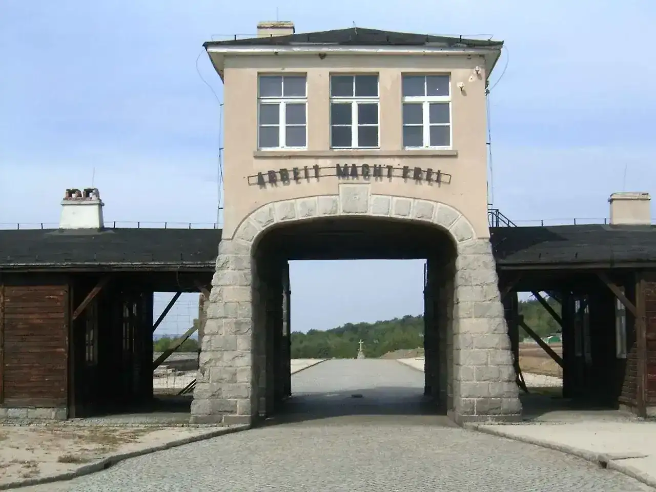 Brama obozu Gross-Rosen z napisem "Arbeit Macht Frei". W oddali krzyż.