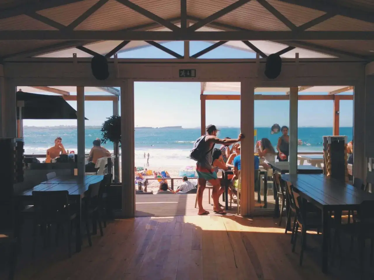 Widok z wnętrza restauracji na plażę i morze. W środku ludzie siedzą przy stołach, na zewnątrz plażowicze. To najlepsza restauracja na Helu.