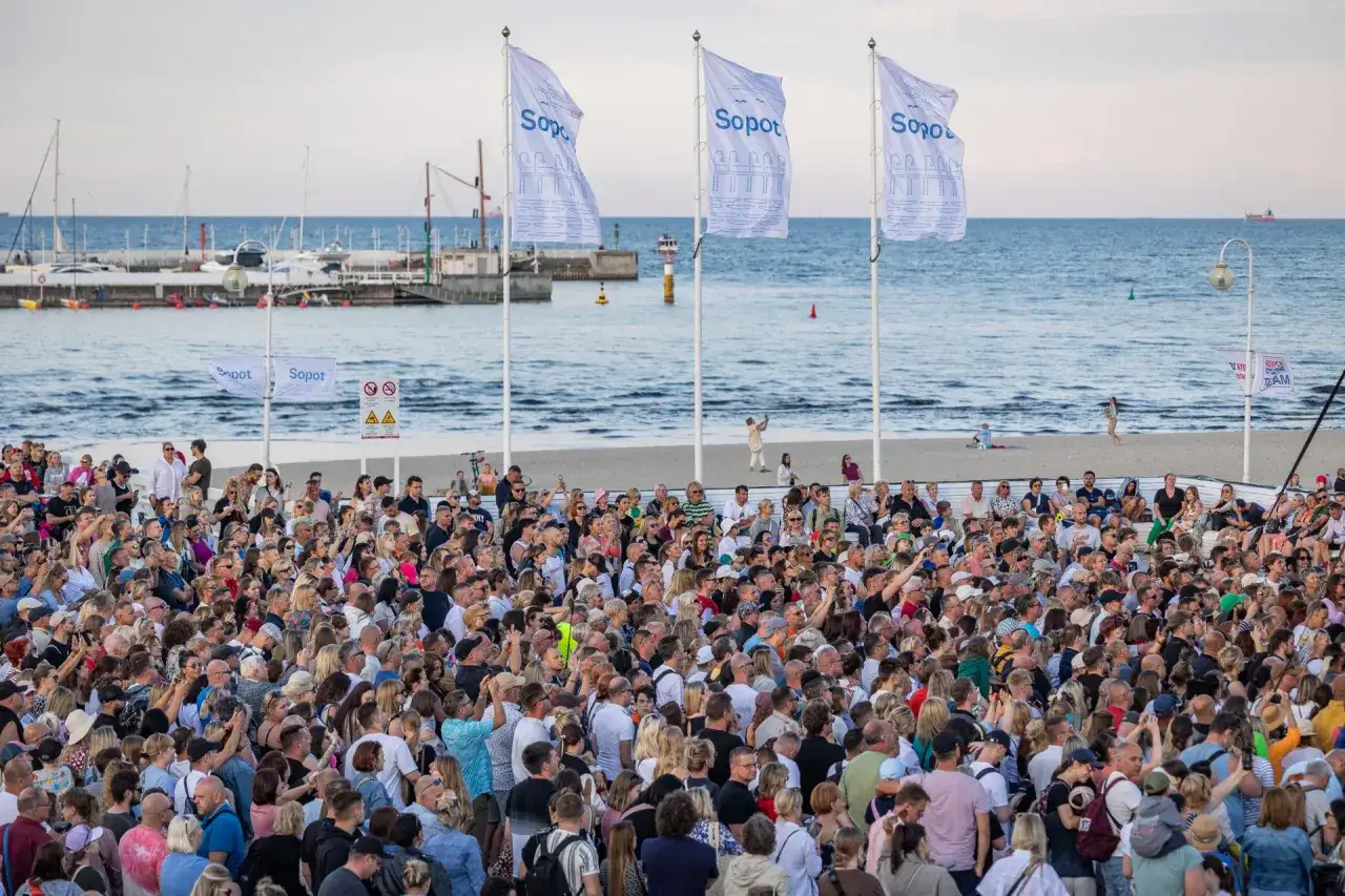 Tłum ludzi na plaży w Sopocie, cieszący się letnimi imprezami. W tle morze i port.