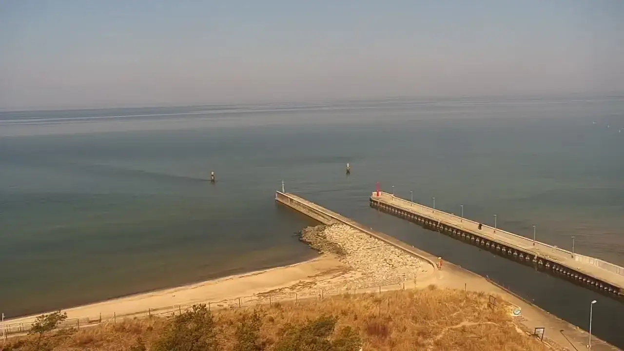 Spokojna plaża z piaszczystym brzegiem i dwoma molo, jedno z czerwoną latarnią. W oddali widać boje nawigacyjne.