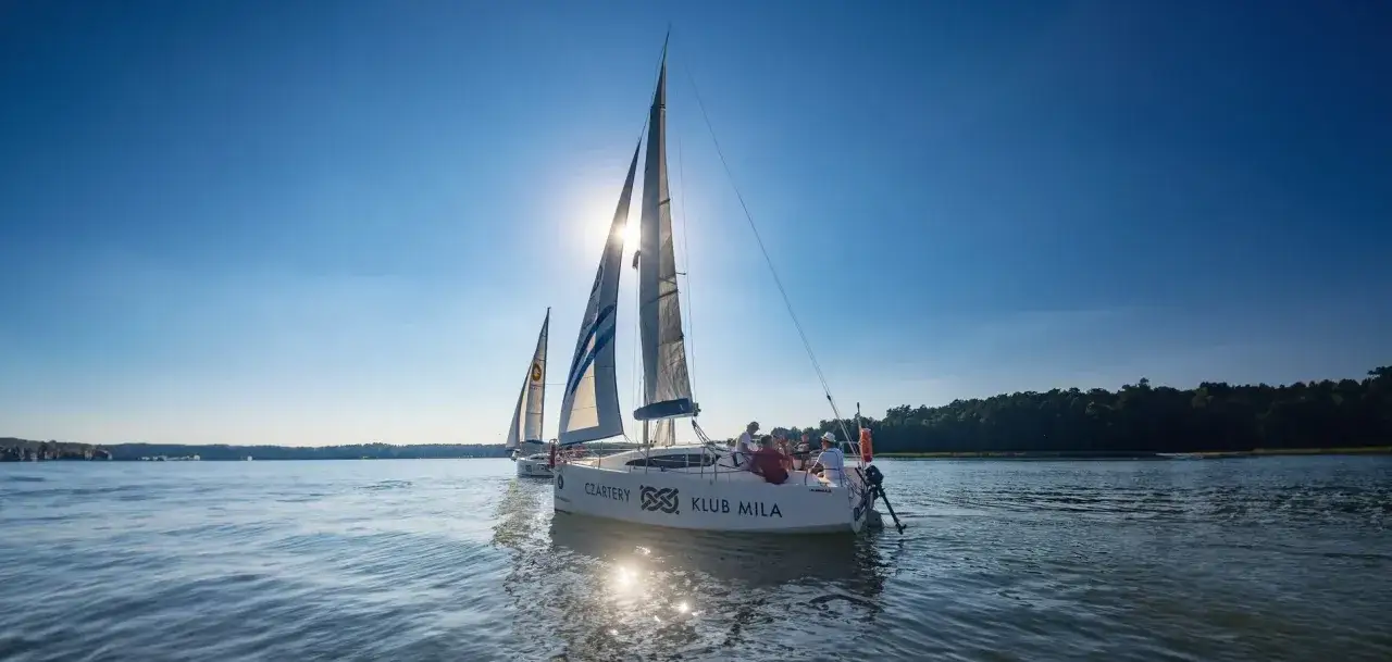 Słońce przebija się przez żagle, zapraszając na wspaniały weekend na Mazurach. Dwie białe łodzie żaglowe płyną po spokojnej wodzie, a na pokładzie widać ludzi cieszących się rejsem.
