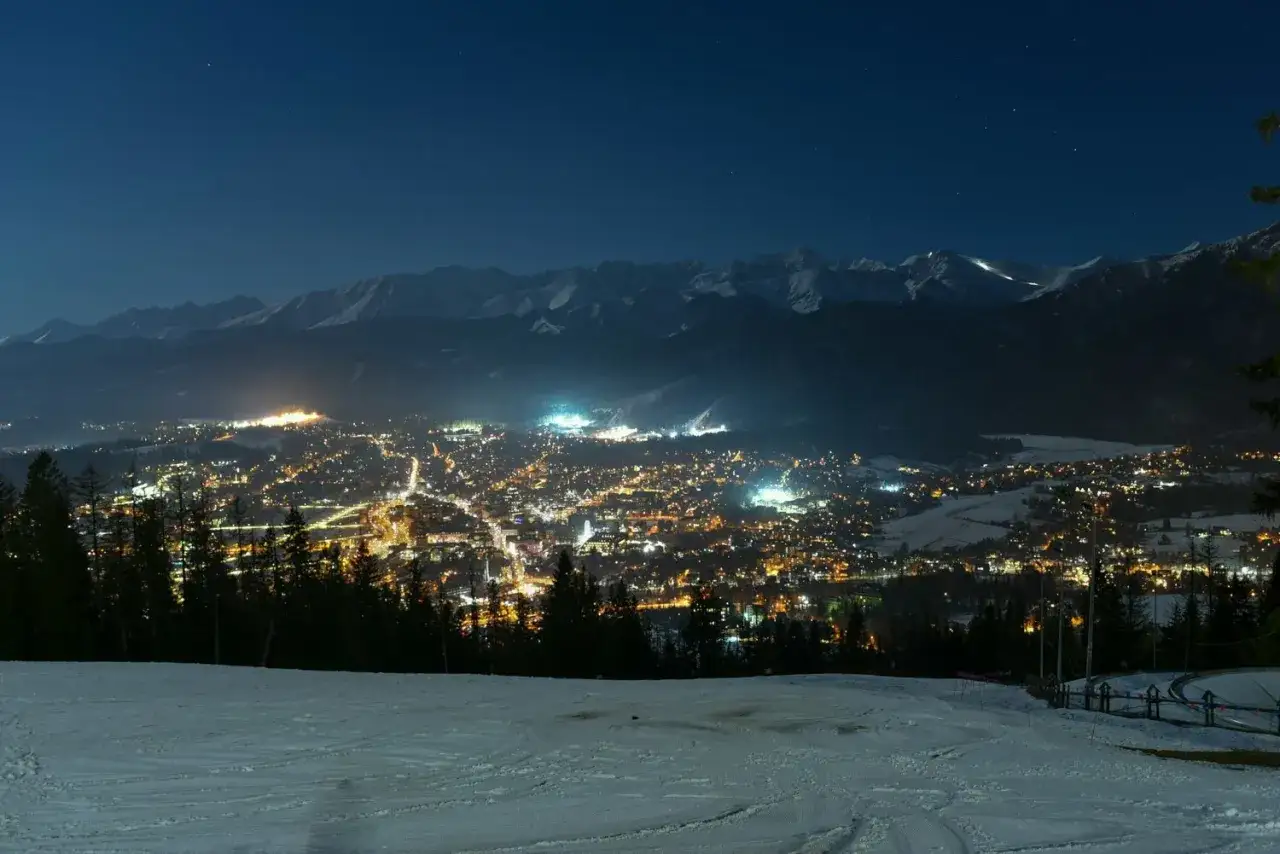 Widok na oświetlone nocą Zakopane z ośnieżonego stoku narciarskiego, w tle góry.