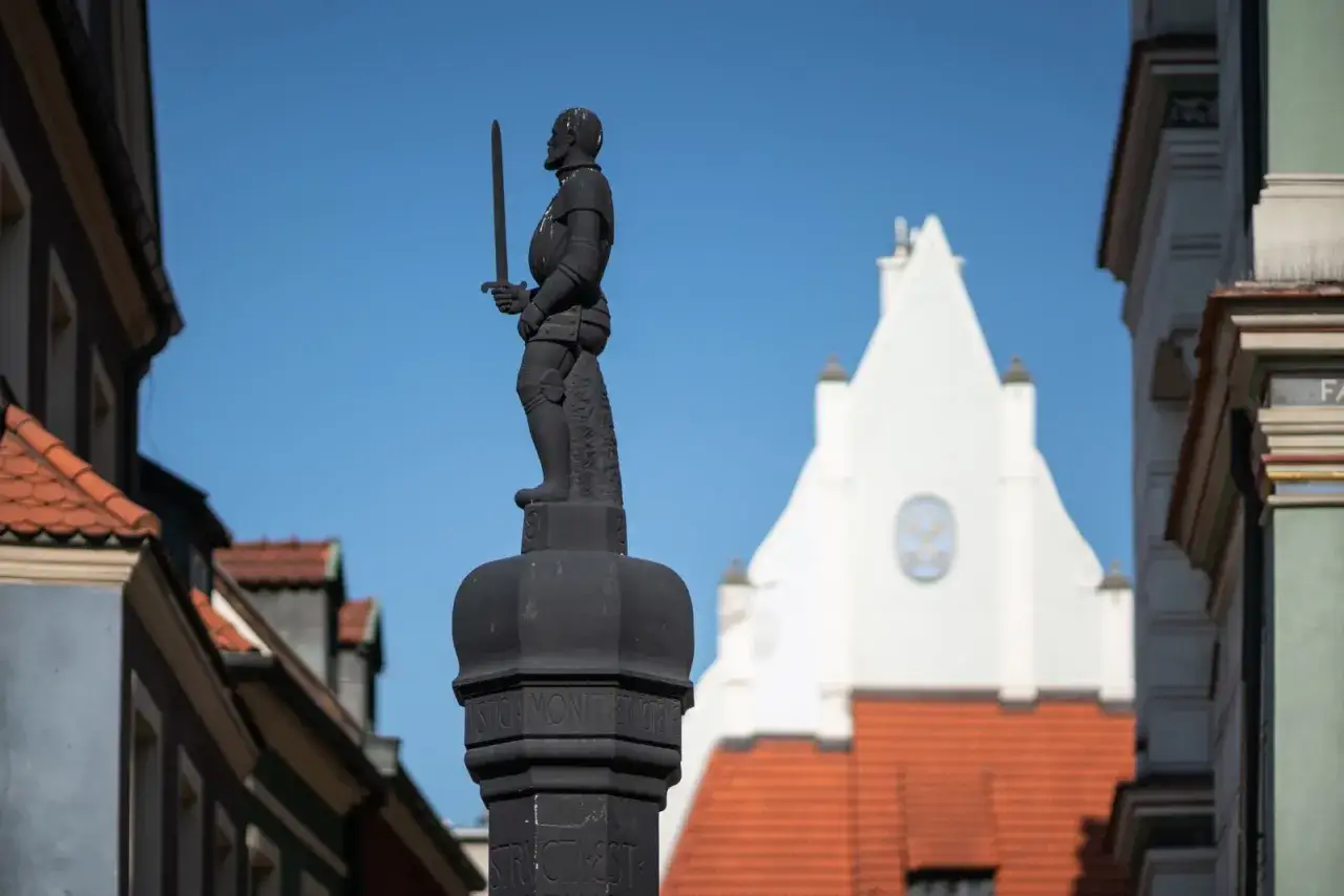 Pręgierz, symbol sprawiedliwości, w postaci rycerza z mieczem na tle zabytkowych kamienic i białej wieży.