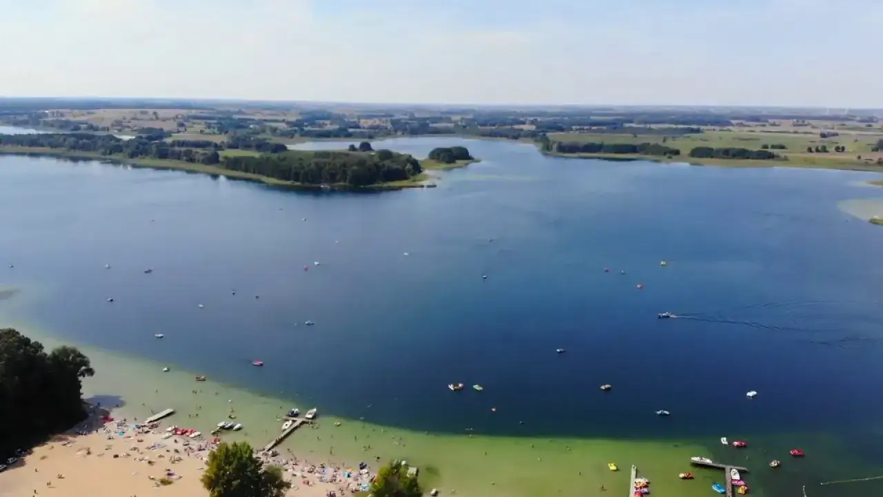 Skorzęcin atrakcje: plaża pełna ludzi, łodzie i rowery wodne na jeziorze, zielone brzegi i wyspy.