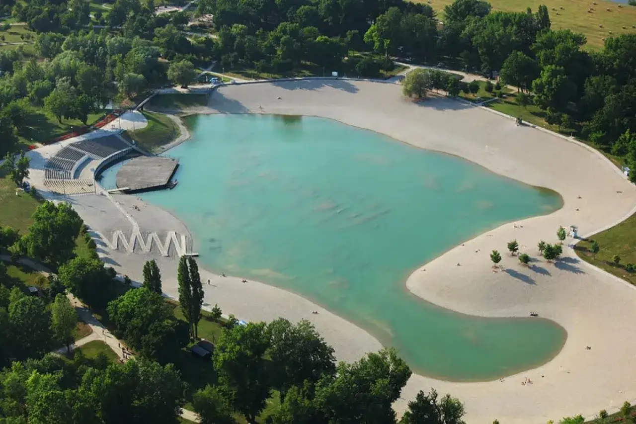 Piękna zagrzeb plaża z turkusową wodą i piaszczystymi brzegami, otoczona zielenią. Widoczne amfiteatr i drewniany pomost.