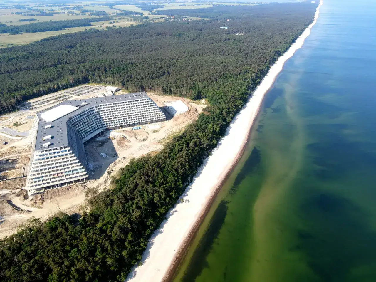 Nowy hotel Gołębiewski nad morzem, w otoczeniu lasu sosnowego, z widokiem na piaszczystą plażę i turkusową wodę.