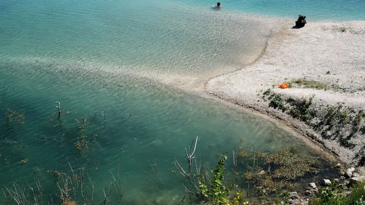 Osoba pływa w krystalicznie czystej wodzie, obok piaszczysta plaża z plecakiem i pomarańczowym ręcznikiem.
