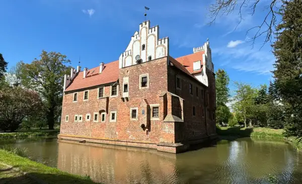 Zamek w Wojnowicach otoczony fosą, z ceglaną fasadą i czerwoną dachówką, pod błękitnym niebem.