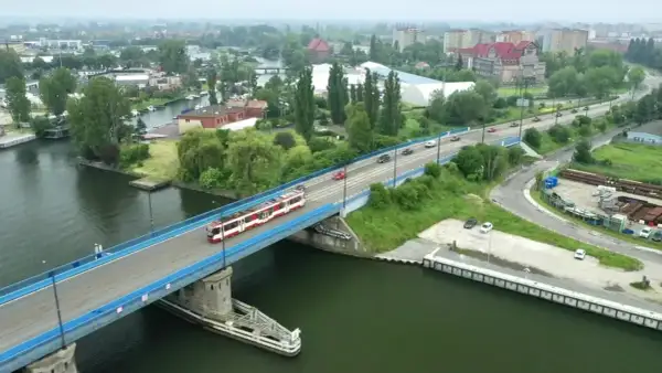Tram z czerwonymi pasami jedzie przez most nad rzeką, w tle widać miasto i tereny przemysłowe, jak na plaży Stogi.