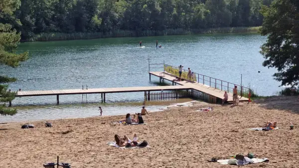Słoneczna borówno plaża nad jeziorem, ludzie odpoczywają na piasku i w wodzie, niektórzy pływają na desce SUP.